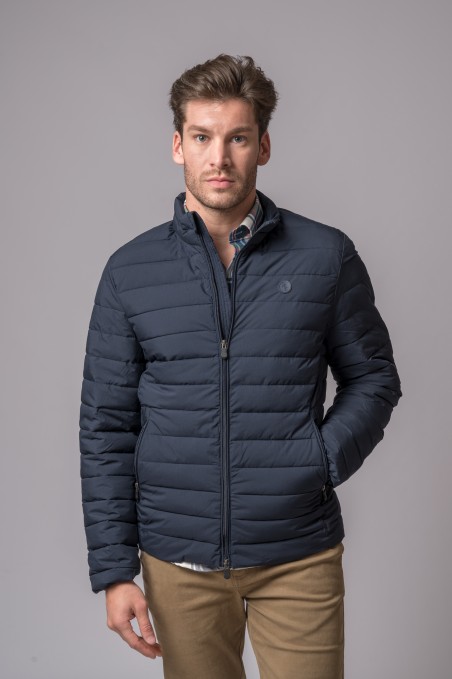 Homem com casaco acolchoado azul escuro e calças bege em fundo cinzento