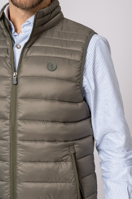 Colete acolchoado verde-oliva com fecho e bolso lateral, usado sobre camisa às riscas azul claro e branco