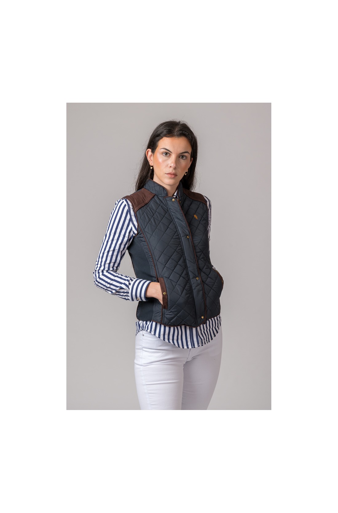 Mulher vestindo colete acolchoado azul escuro com remates castanhos sobre camisa às riscas brancas e azuis