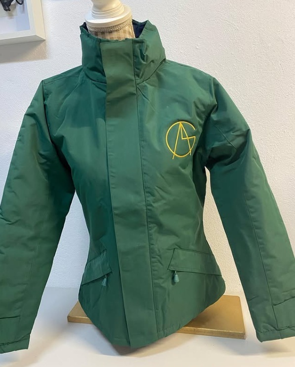 Casaco verde escuro com bordado amarelo 'AG' no peito exposto num manequim