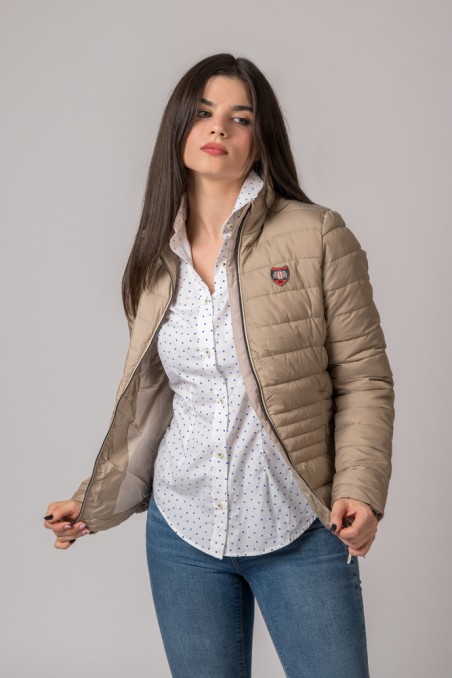 Mulher com casaco bege acolchoado, camisa branca com pequenos pontos e calças de ganga azuis.