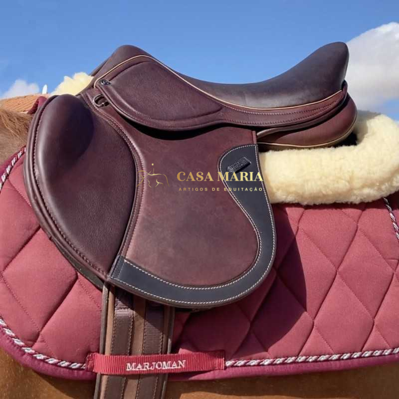 Sela de equitação em couro castanho sobre manta acolchoada vermelha com detalhes brancos e vermelhos e palavras MARJOMAN e CASA MARIA visíveis