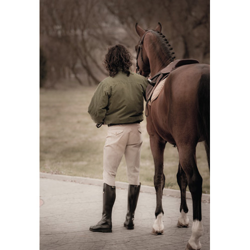 Homem de casaco verde e botas pretas lado a lado com cavalo castanho com crina trançada ao ar livre