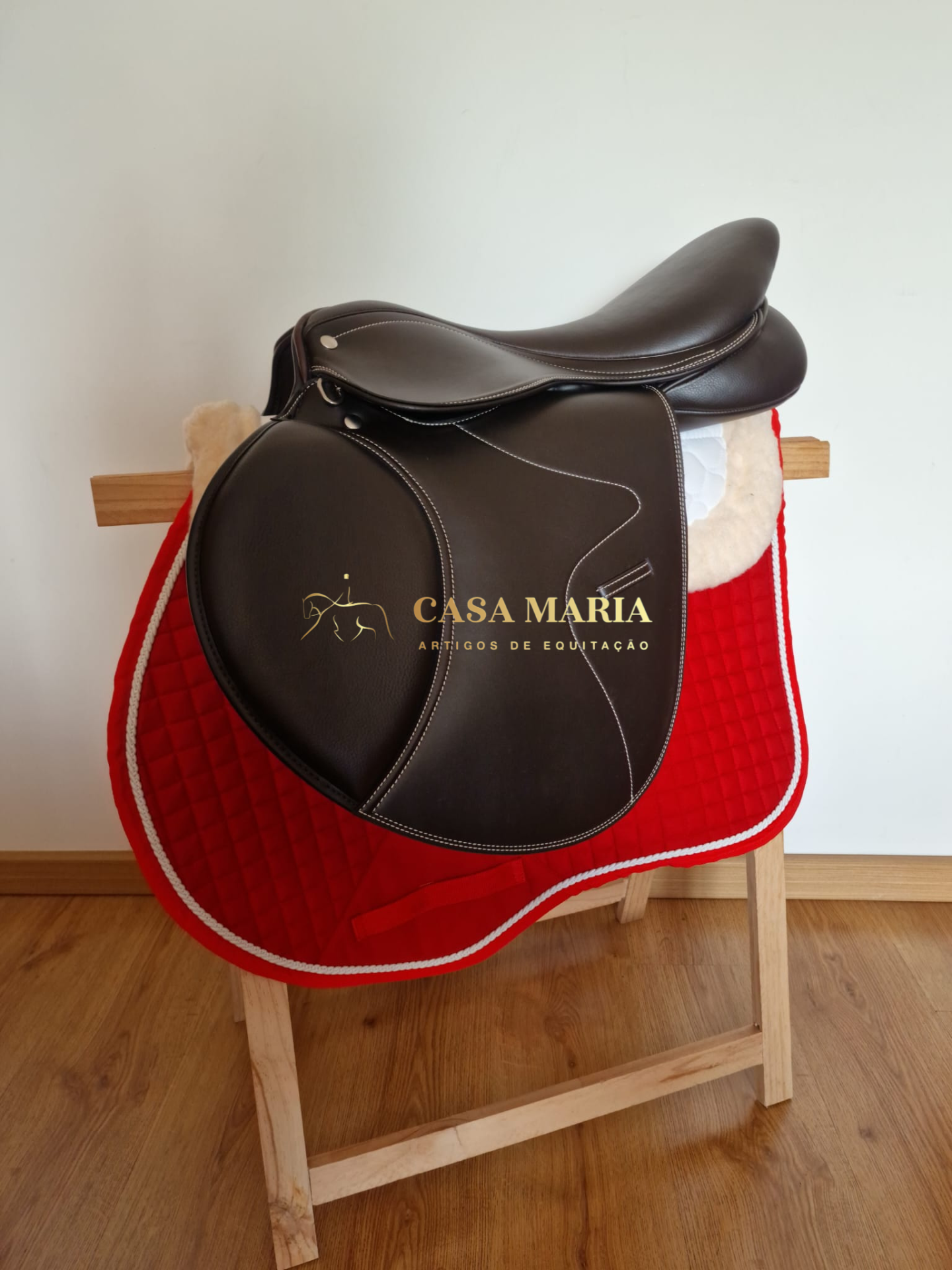 Sela de cavalo em pele castanha escura sobre suporte de madeira com manta de sela vermelha acolchoada