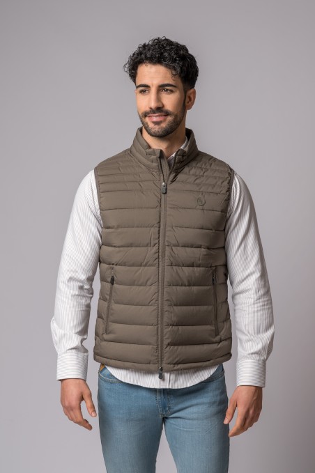 Colete acolchoado castanho claro em homem com camisa branca e calças de ganga, fundo cinza.