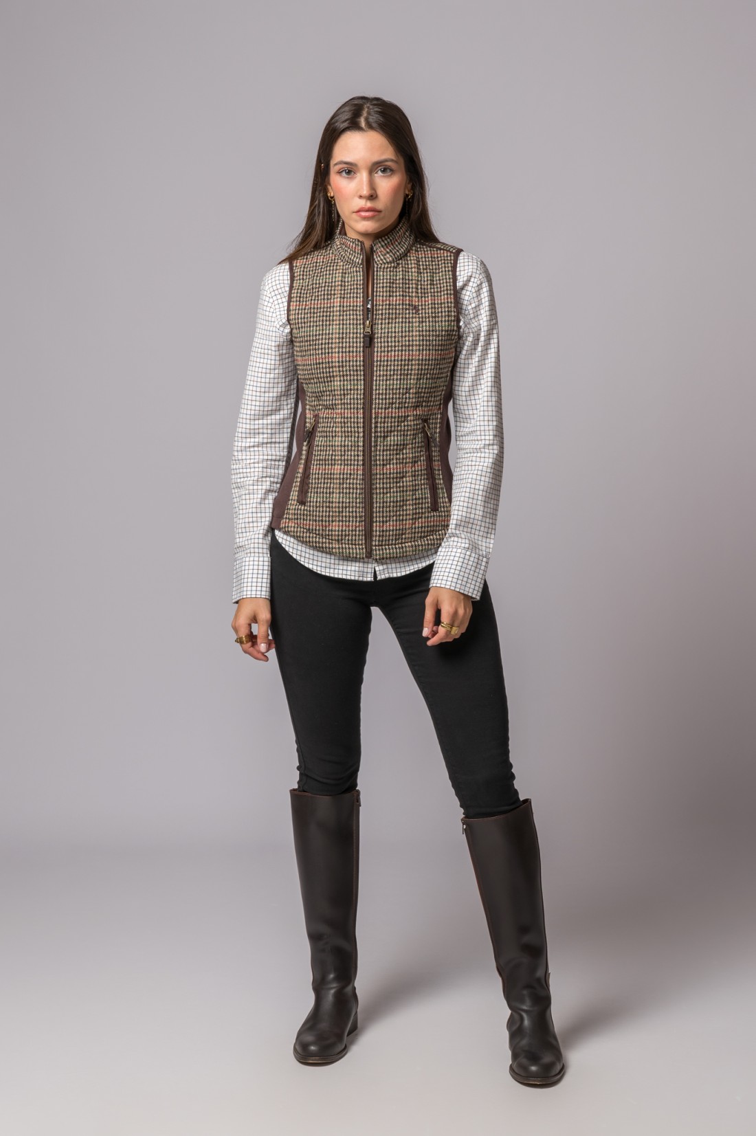 Mulher a vestir colete castanho com padrão pied-de-poule, camisa quadriculada branca, calças pretas e botas castanhas em fundo cinza.