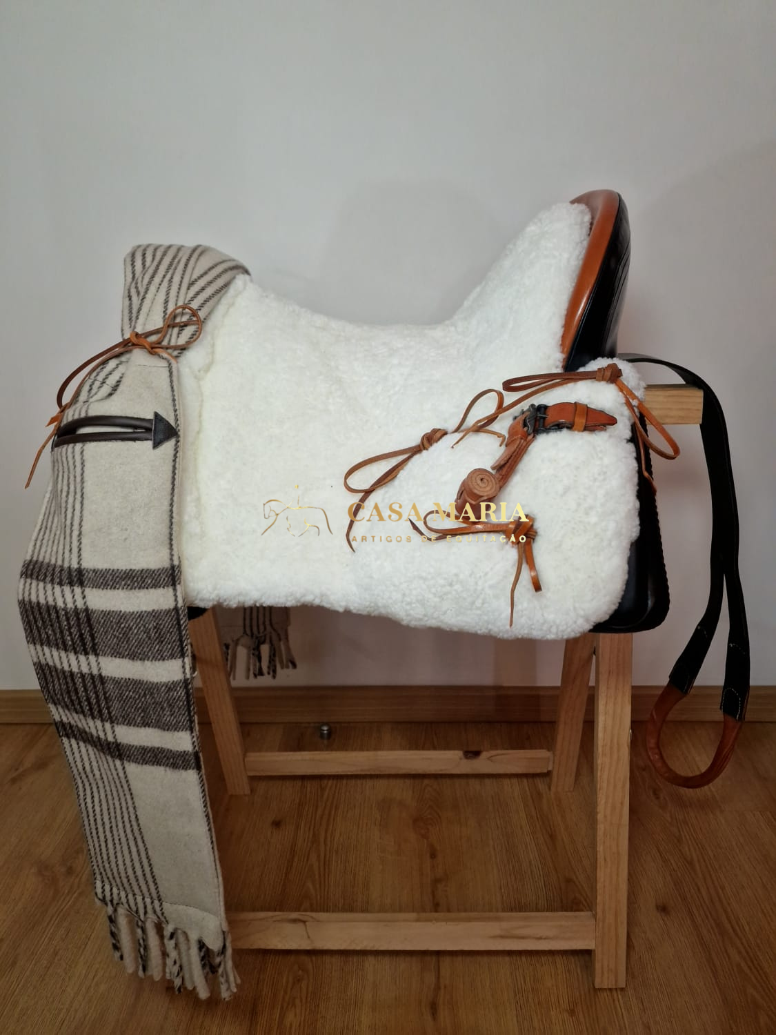 Sela de equitação branca com detalhes castanhos e manta às riscas num suporte de madeira
