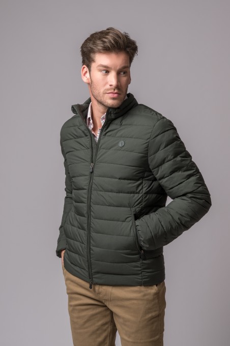 Homem com casaco acolchoado verde-escuro e calças castanhas frente a fundo cinzento
