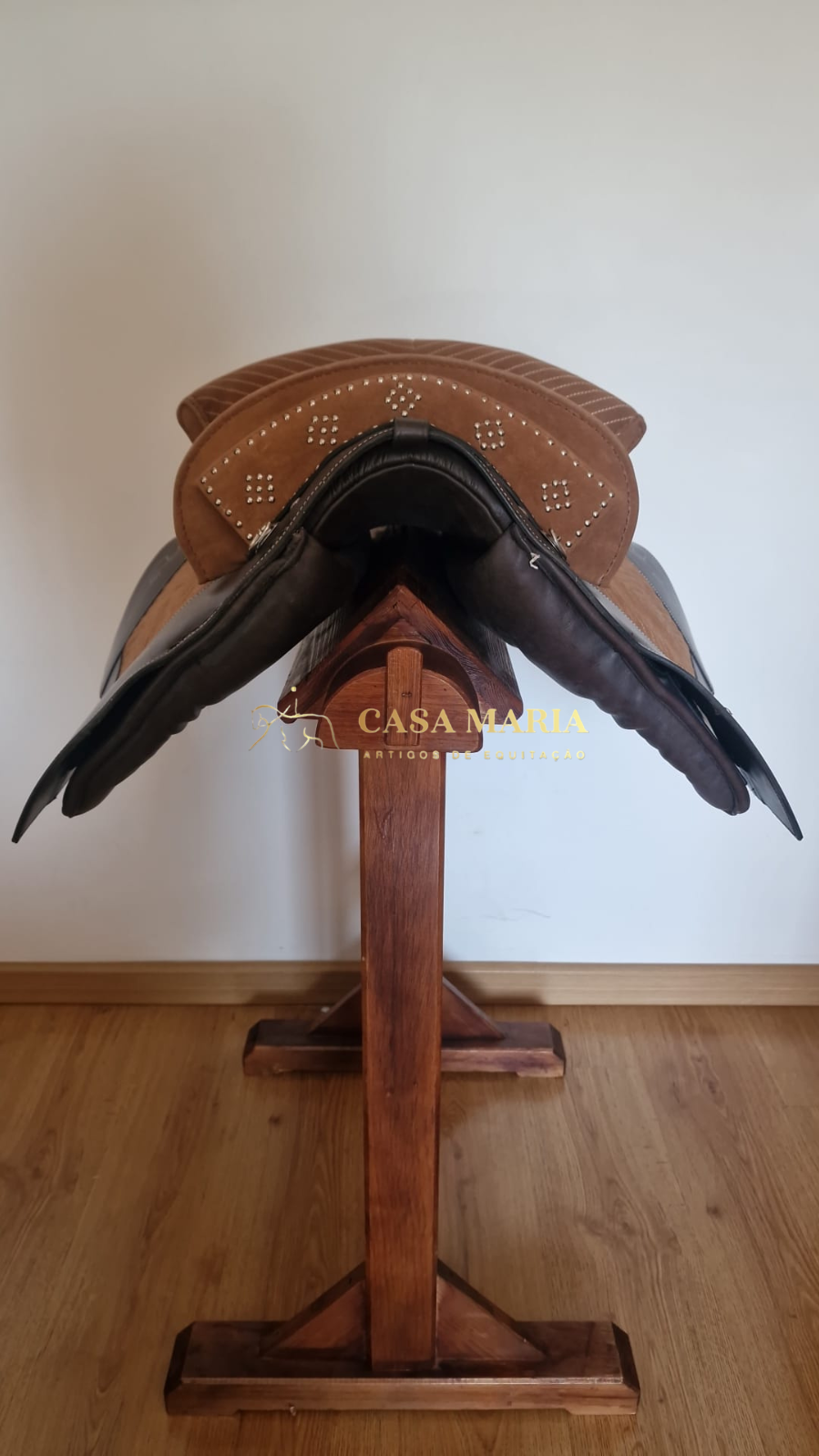 Sela de equitação castanha com detalhes perfurados em padrão geométrico apoiada em suporte de madeira