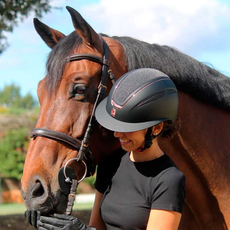 Capacete de equitação preto com detalhes rosa usado por mulher junto a cavalo castanho em exterior