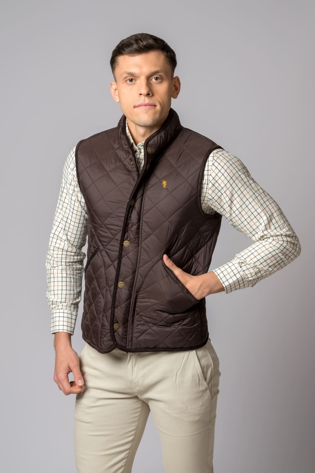 Homem a usar colete acolchoado castanho com camisa xadrez e calças bege