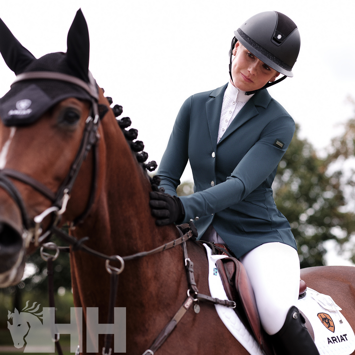 Cavaleira com roupa de equitação azul e branca montada num cavalo castanho com touca preta e manta branca com logótipos