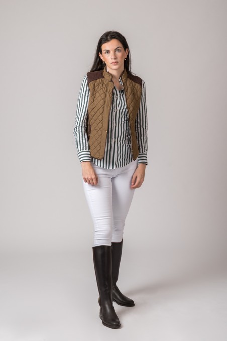 Mulher com camisa riscada, colete castanho acolchoado, calças brancas e botas pretas em fundo cinzento
