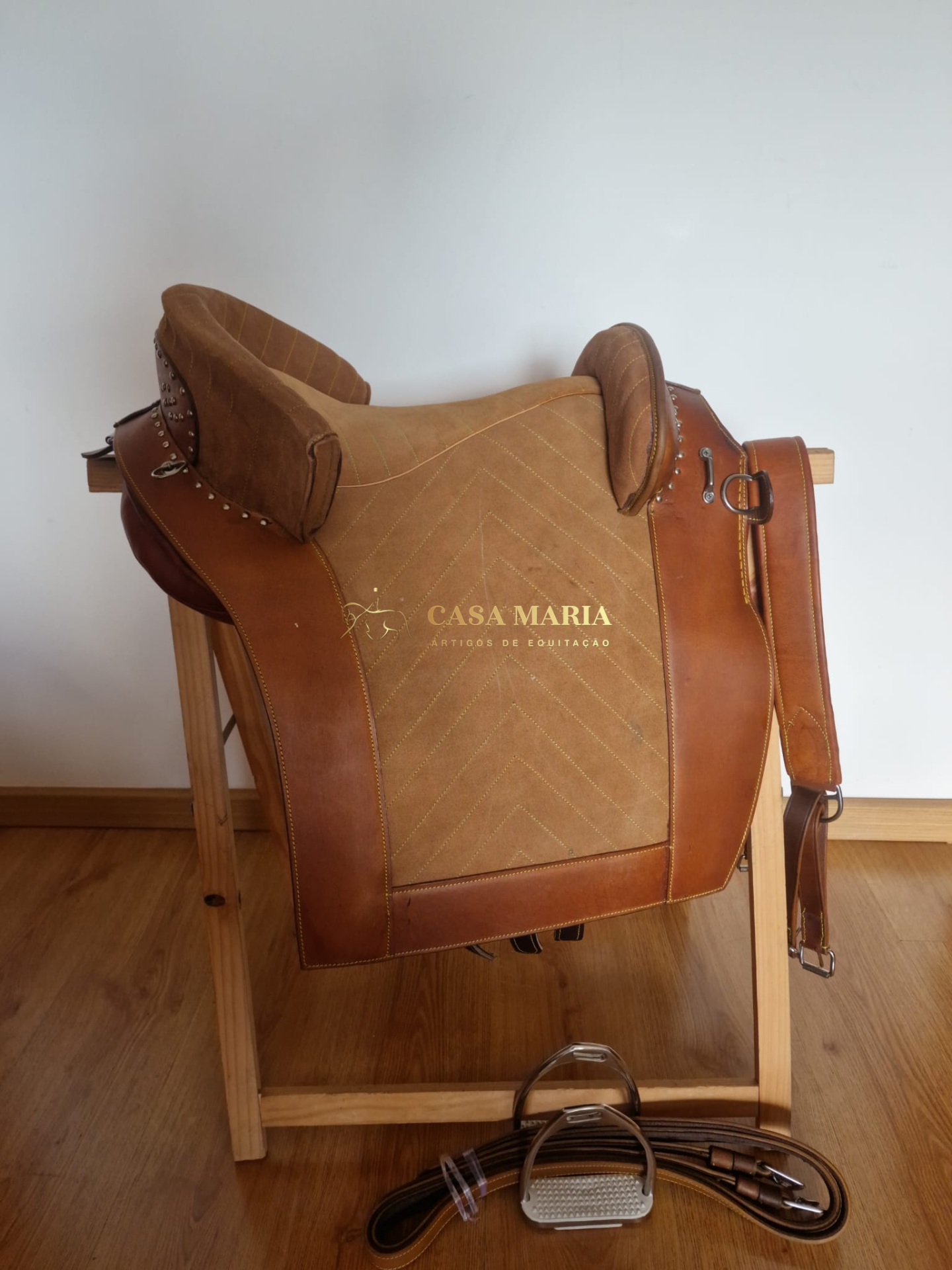 Sela de equitação CASA MARIA em couro castanho sobre suporte de madeira com estribo e rédea no chão.