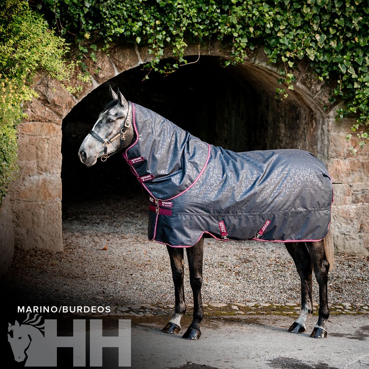 Cobertura azul marinho para cavalo com detalhes bordeaux, montada num cavalo em frente a uma parede de pedra