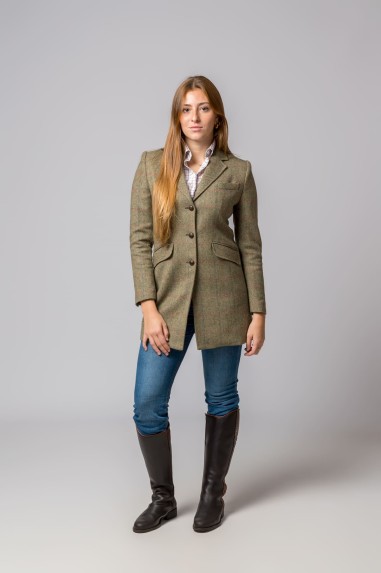 Modelo a usar casaco comprido tweed castanho-verde com botões, calças jeans azuis e botas pretas, fundo cinza.