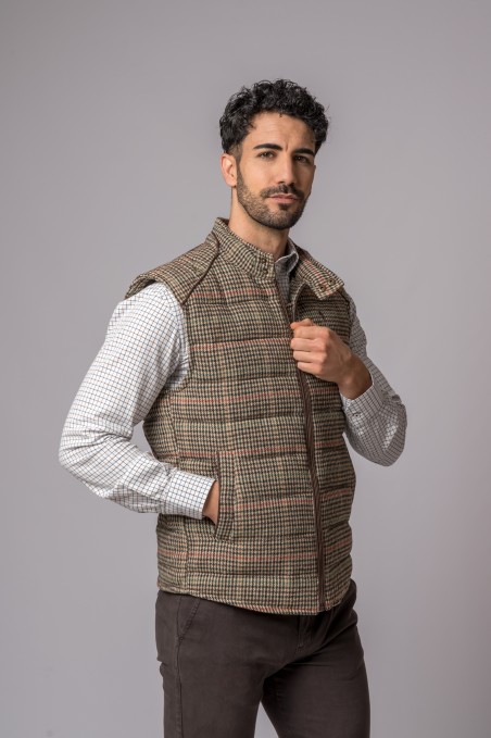 Homem com colete xadrez castanho e bege, camisa branca xadrez pequena e calças castanhas escuras
