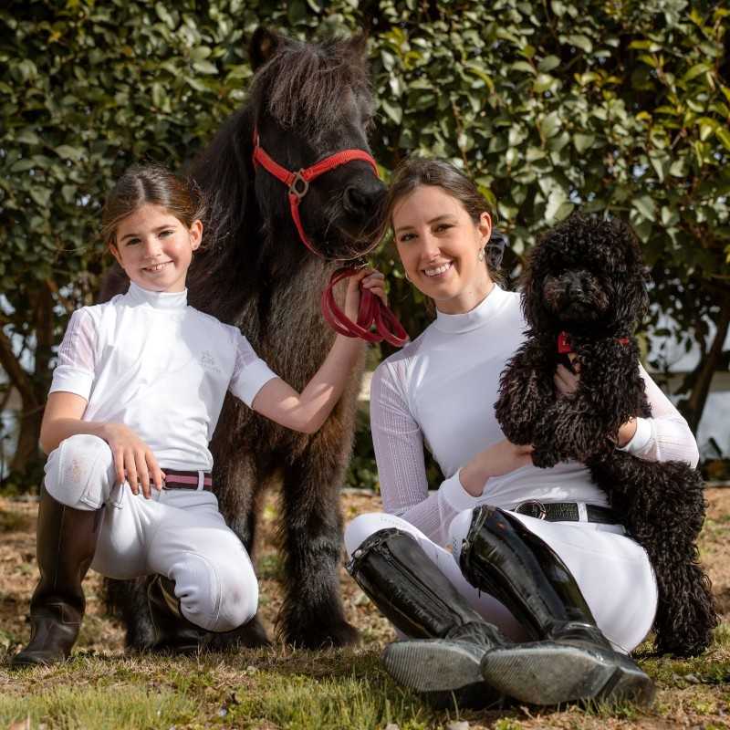 Duas pessoas com roupa branca de equitação, égua preta com peitoral vermelho e cão preto ao ar livre.