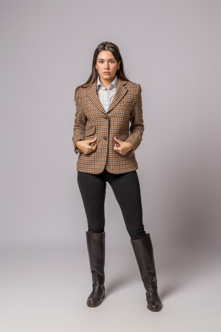 Mulher a usar blazer xadrez castanho com camisa branca, calças pretas e botas pretas em fundo cinzento.