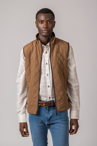 Homem com colete matelassê castanho, camisa xadrez, jeans azuis e cinto, fundo cinza