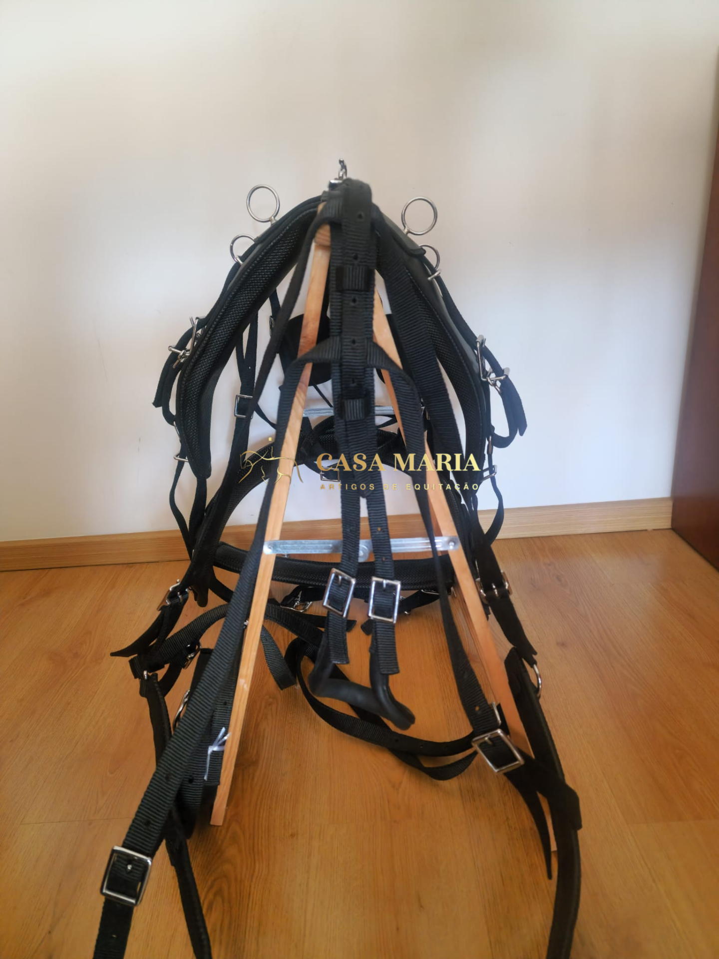 Arnês preto com fivelas metálicas e argolas sobre estrutura de madeira em chão de madeira