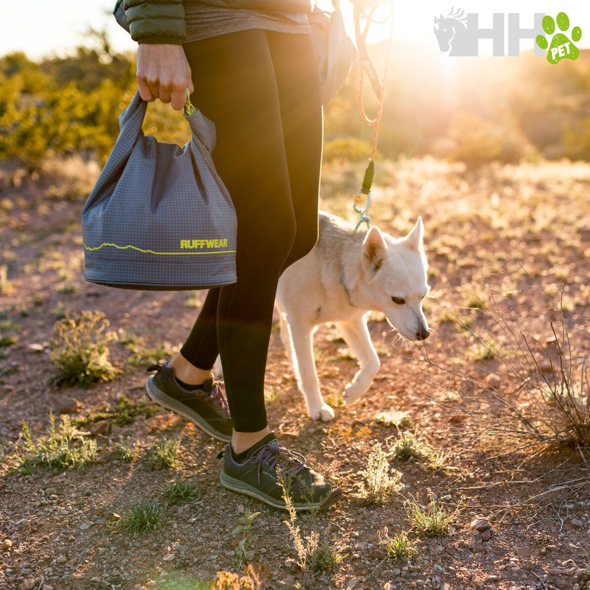Pessoa a caminhar com mochila cinzenta RUFFWEAR e cão branco em trilho