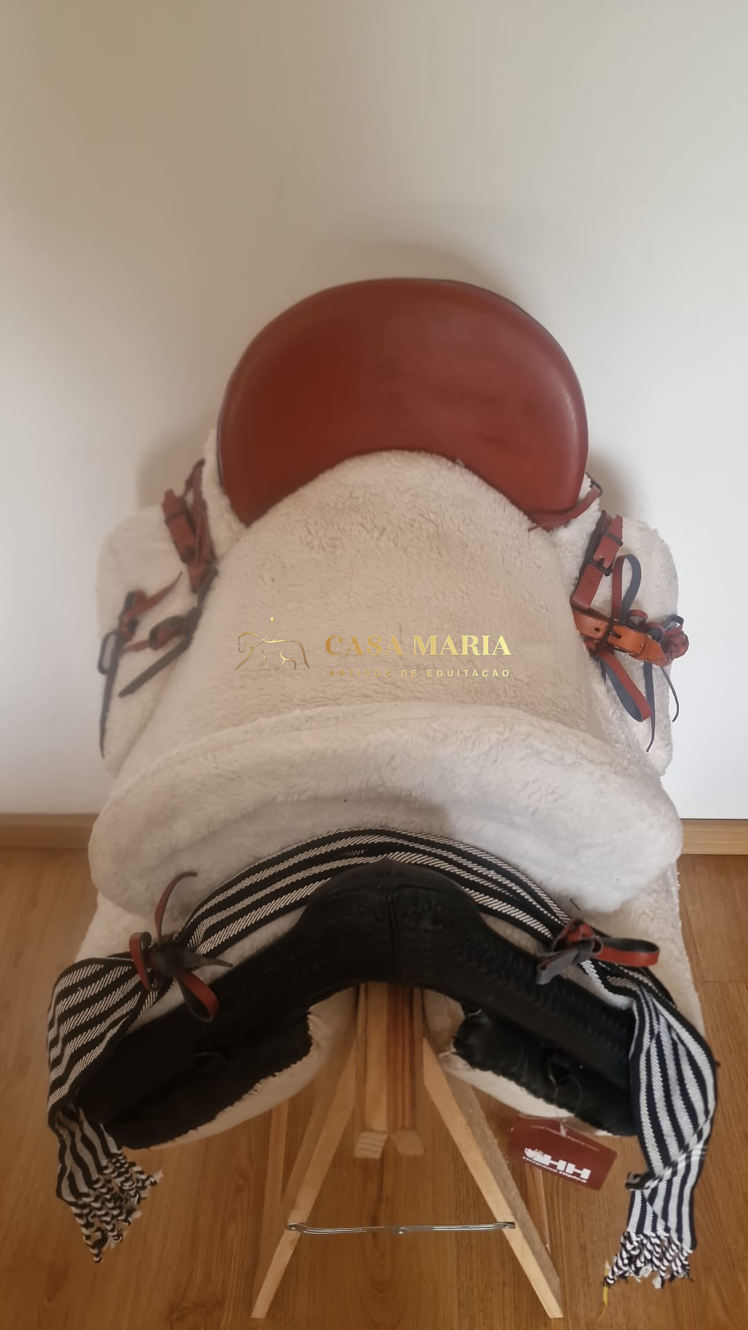 Sela de equitação castanha com base branca e manta listrada preta e branca sobre suporte de madeira