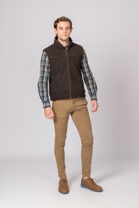 Homem vestindo colete castanho escuro, camisa xadrez, calças bege e sapatos castanhos com fundo cinza