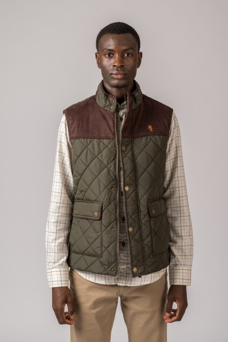 Colete masculino verde-oliva acolchoado com parte superior castanha e bolsos frontais, usado sobre camisa xadrez e calças castanhas.