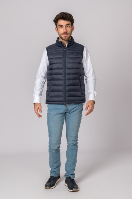 Homem com colete acolchoado azul escuro, camisa branca, jeans e sapatilhas escuras