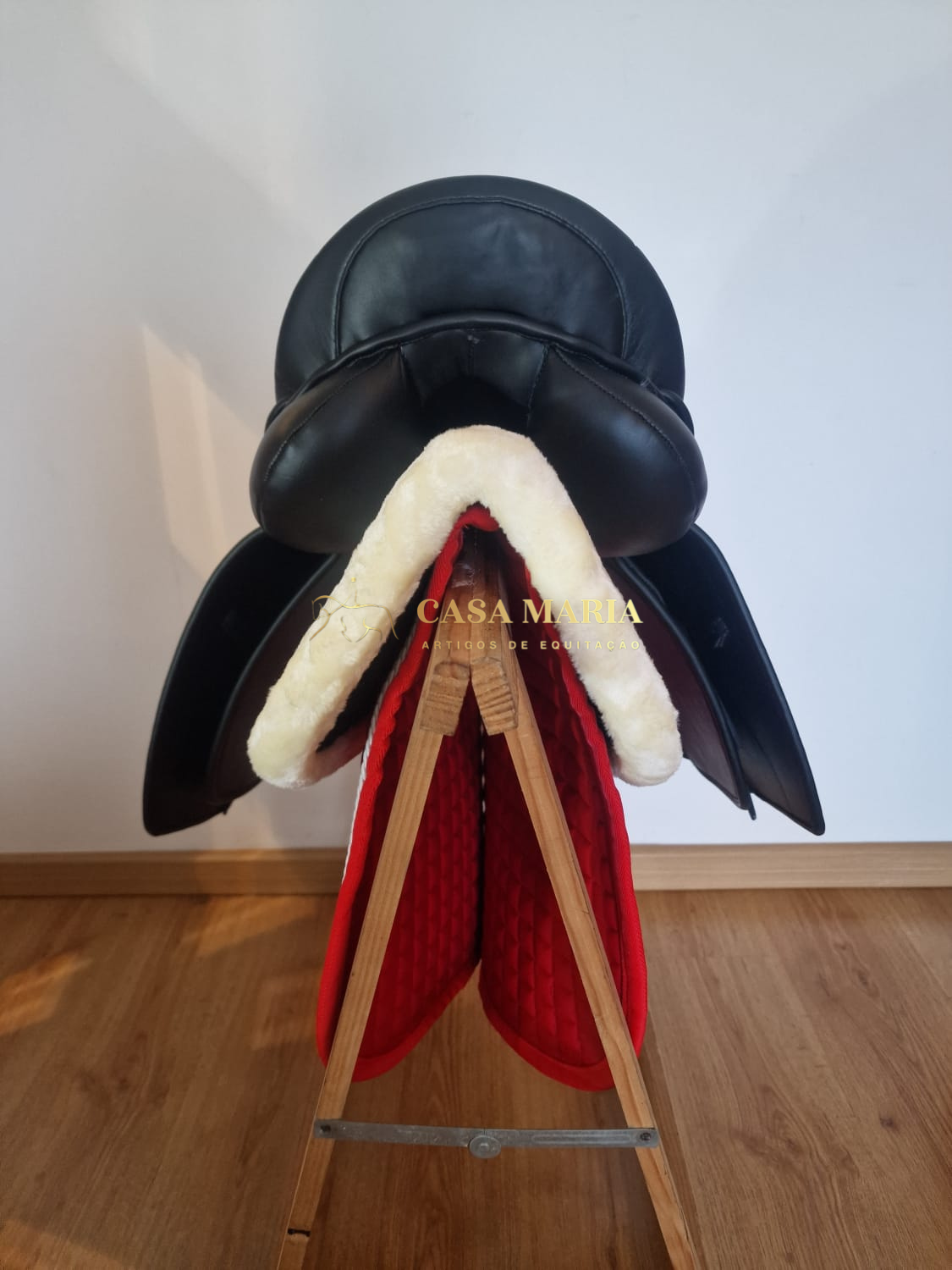 Sela de equitação preta com revestimento branco e manta vermelha sobre suporte de madeira