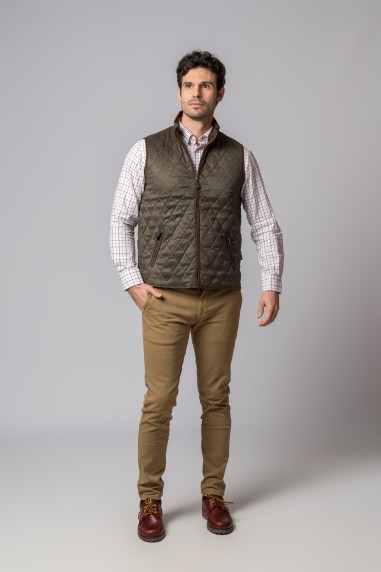 Homem com colete verde-oliva acolchoado, camisa xadrez, calças castanhas e sapatos castanhos num fundo cinza.