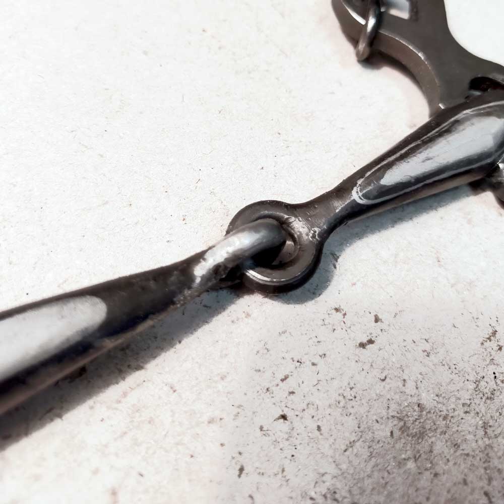 Peça metálica castanho escuro com manchas prateadas e ligação circular sobre superfície branca suja.