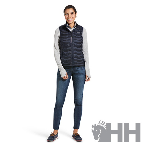 Mulher vestindo colete azul escuro acolchoado com padrão ondulado e zíper frontal, jeans azul e camisola cinzenta, em fundo branco com logo HH cinzento.