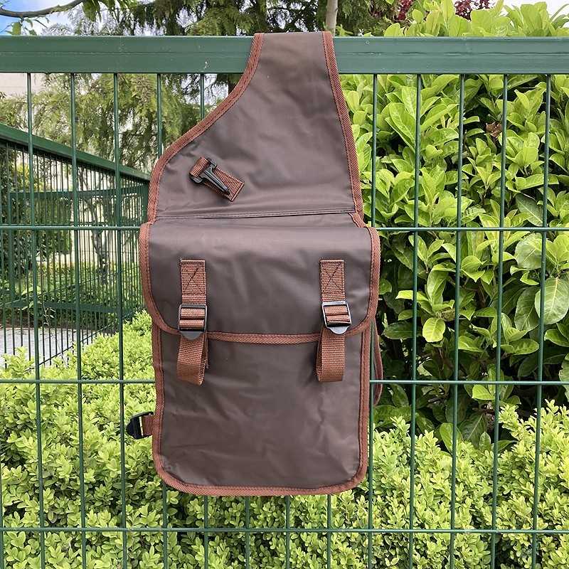 Mochila castanha escura com alças e fechos castanhos pendurada numa vedação verde com plantas ao fundo