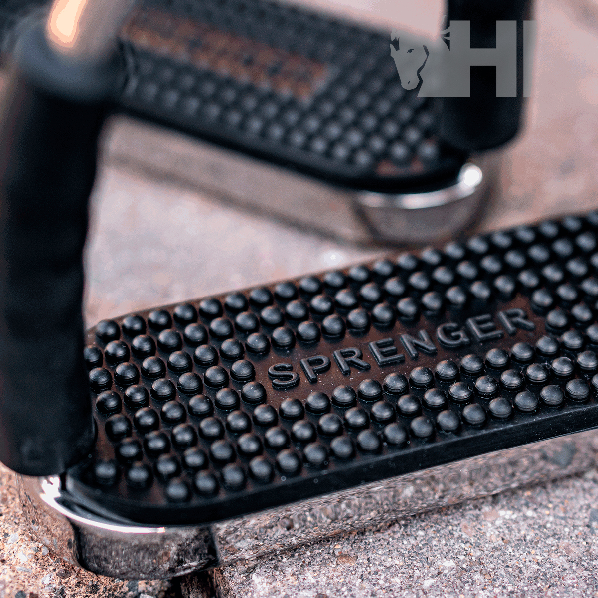 Estribos metálicos com apoio de borracha texturizada preta e texto SPRNGER sobre piso de pedra
