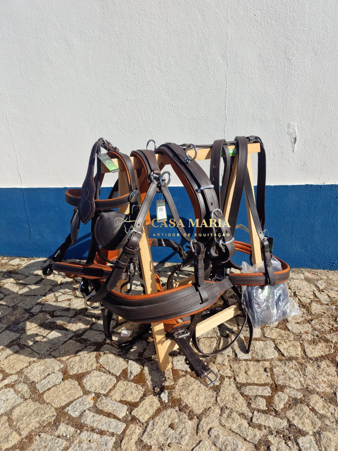 Arnês de equitação castanho pendurado em suporte de madeira num chão de pedra perto de uma parede branca com faixa azul