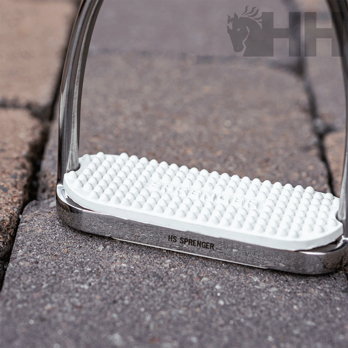 Estribo de equitação metálico com base branca antiderrapante sobre pavimento de blocos.