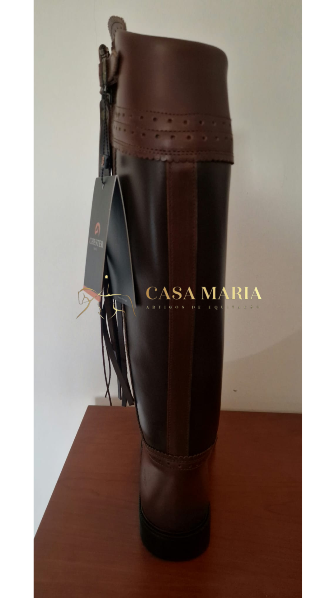 Bota alta castanha de equitação com etiqueta da marca à frente sobre mesa de madeira.