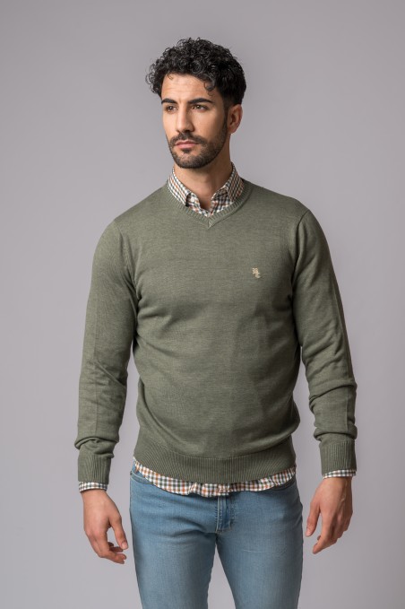 Homem com camisola verde, camisa xadrez e jeans azul claro em fundo cinza.