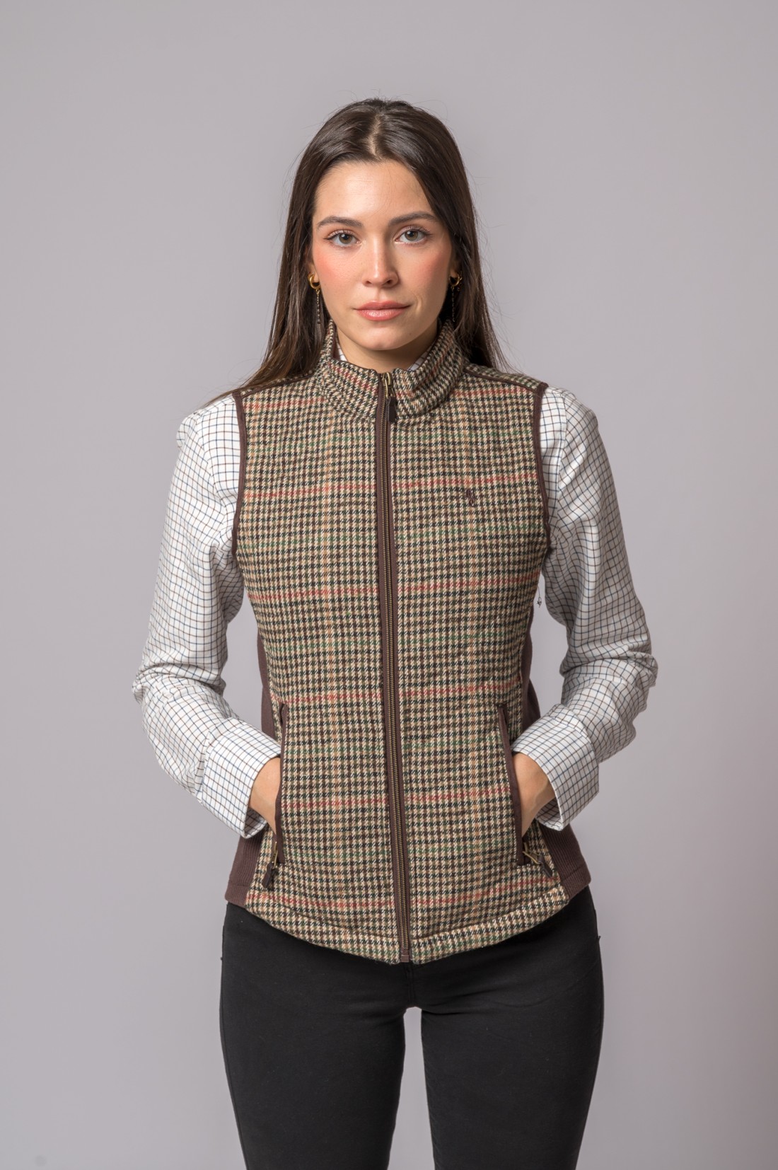 Colete xadrez castanho bege e vermelho com gola alta e fecho de correr, usado por mulher com camisa branca quadriculada e calças pretas.
