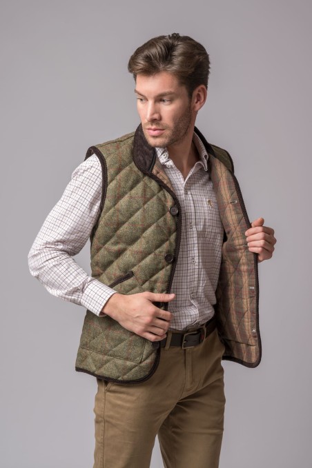 Homem a vestir colete matelassado verde com detalhes castanhos escuros e camisa bege com padrão quadrado