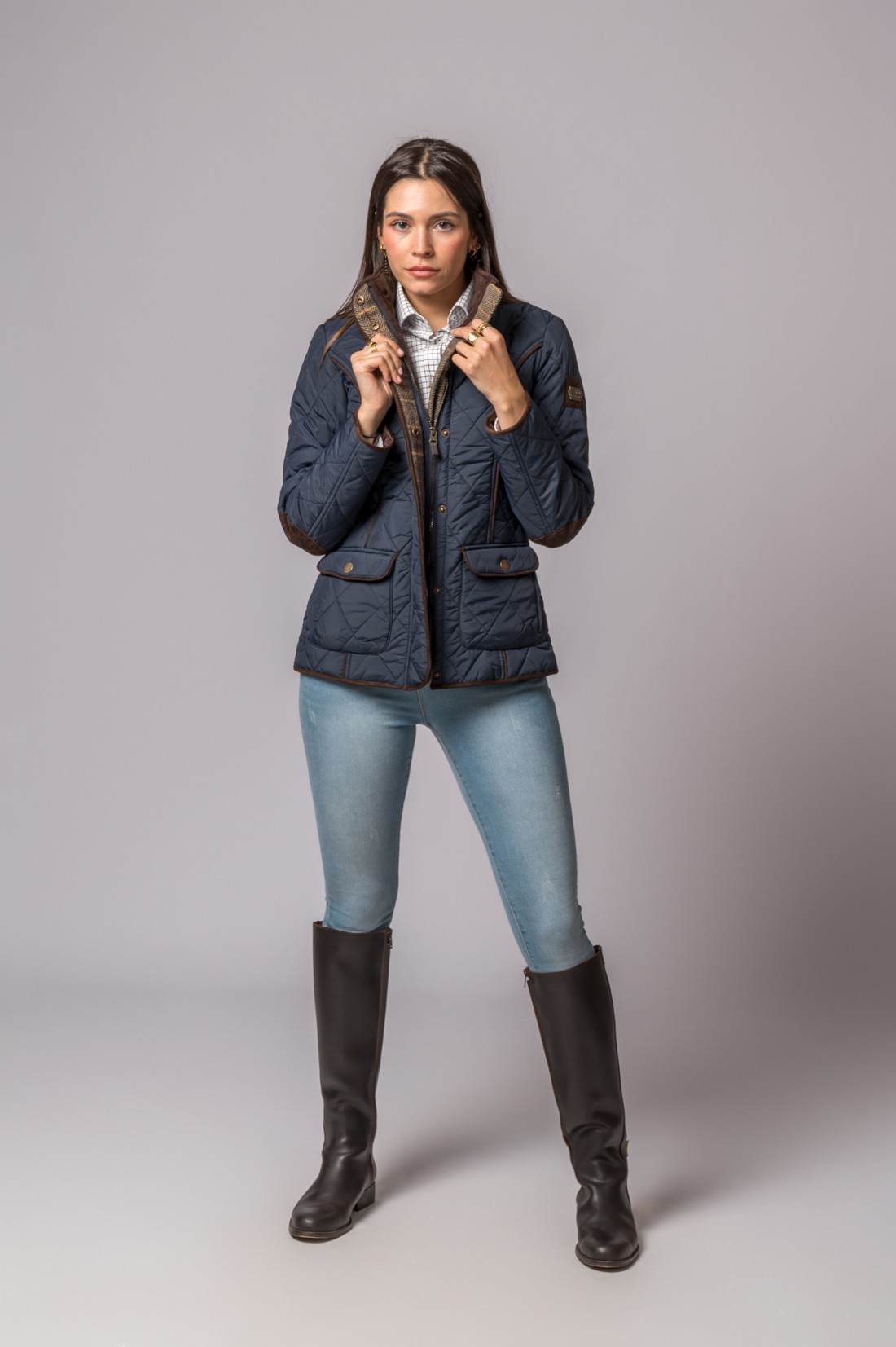 Mulher a vestir casaco acolchoado azul escuro, jeans e botas altas pretas, fundo cinzento.