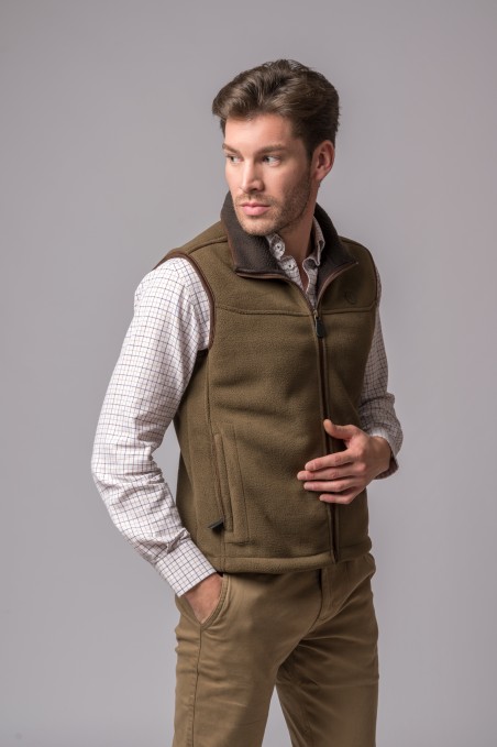 Homem com colete castanho polar, camisa branca xadrez e calças castanho claro em fundo cinzento