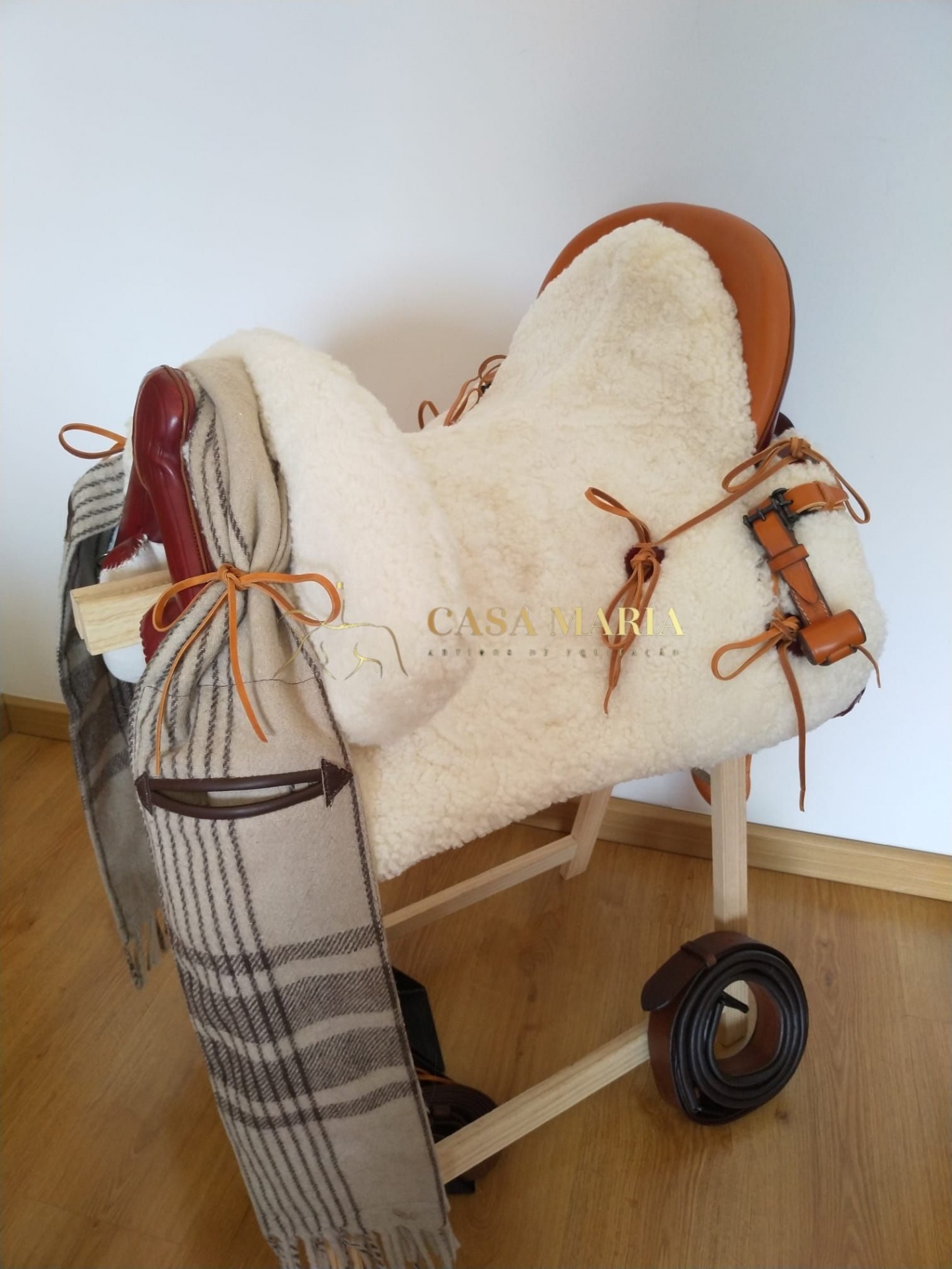Sela de equitação em couro castanho claro com tecido felpudo branco numa estrutura de madeira, com cinto e tecido xadrez