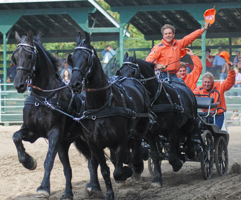 Carroça com cavalos pretos e homens com roupa laranja numa pista coberta