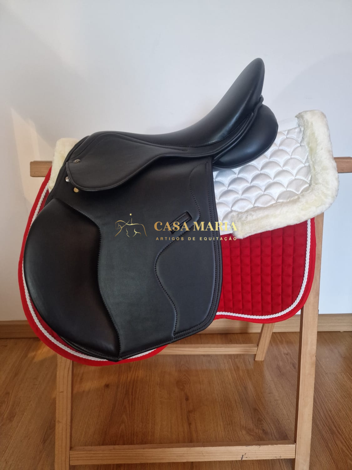 Sela de equitação preta em couro com mantas branca e vermelha sobre suporte de madeira