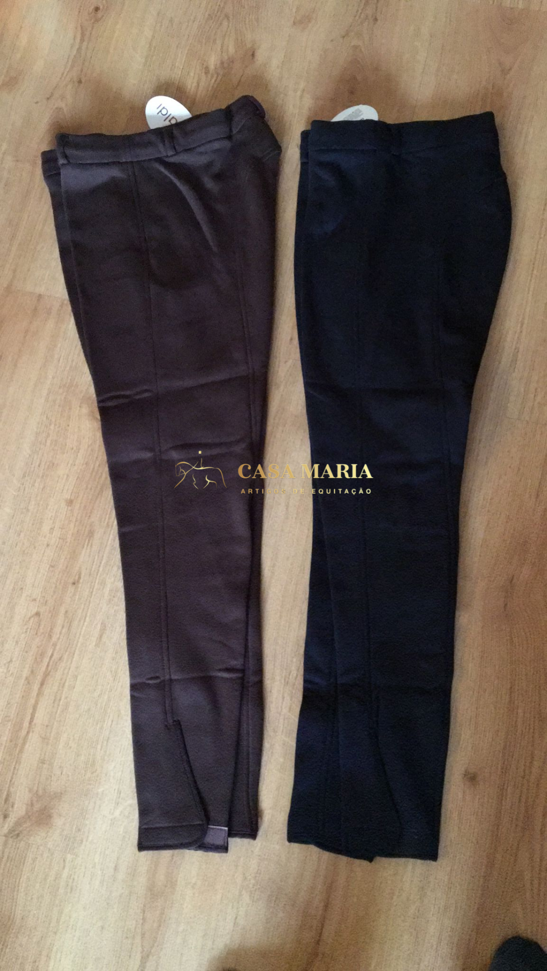 Duas calças de equitação castanha escura e preta, estendidas no chão de madeira