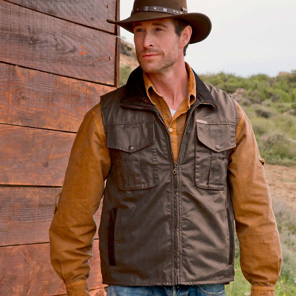 Homem com casaco castanho escuro e chapéu castanho ao ar livre junto a uma parede de madeira.