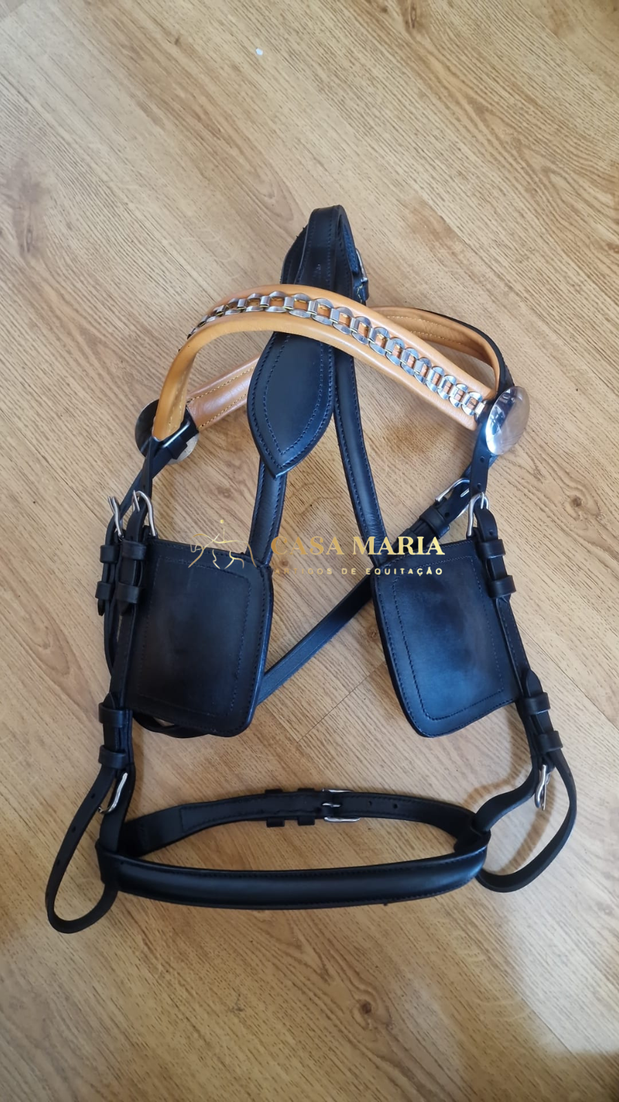 Equipamento equestre em couro preto e bege com detalhes metálicos prateados sobre chão de madeira.